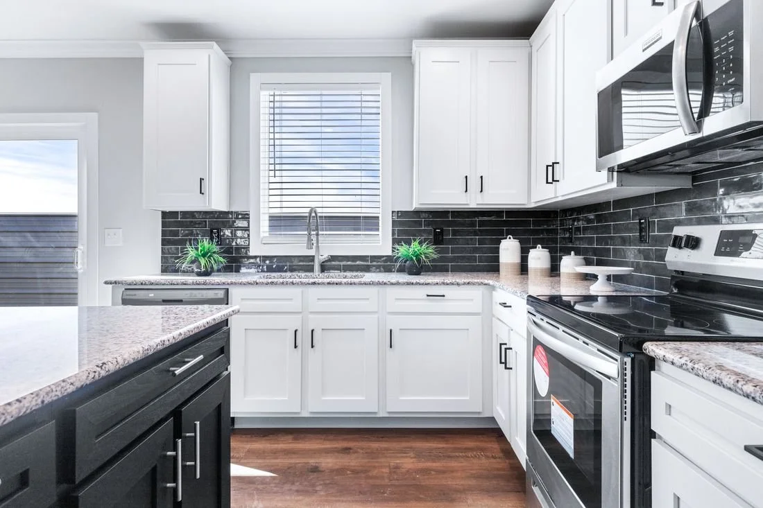 Modern kitchen with white cabinets, black hardware, granite countertops, black backsplash, stainless steel appliances, and a window above the sink with blinds.