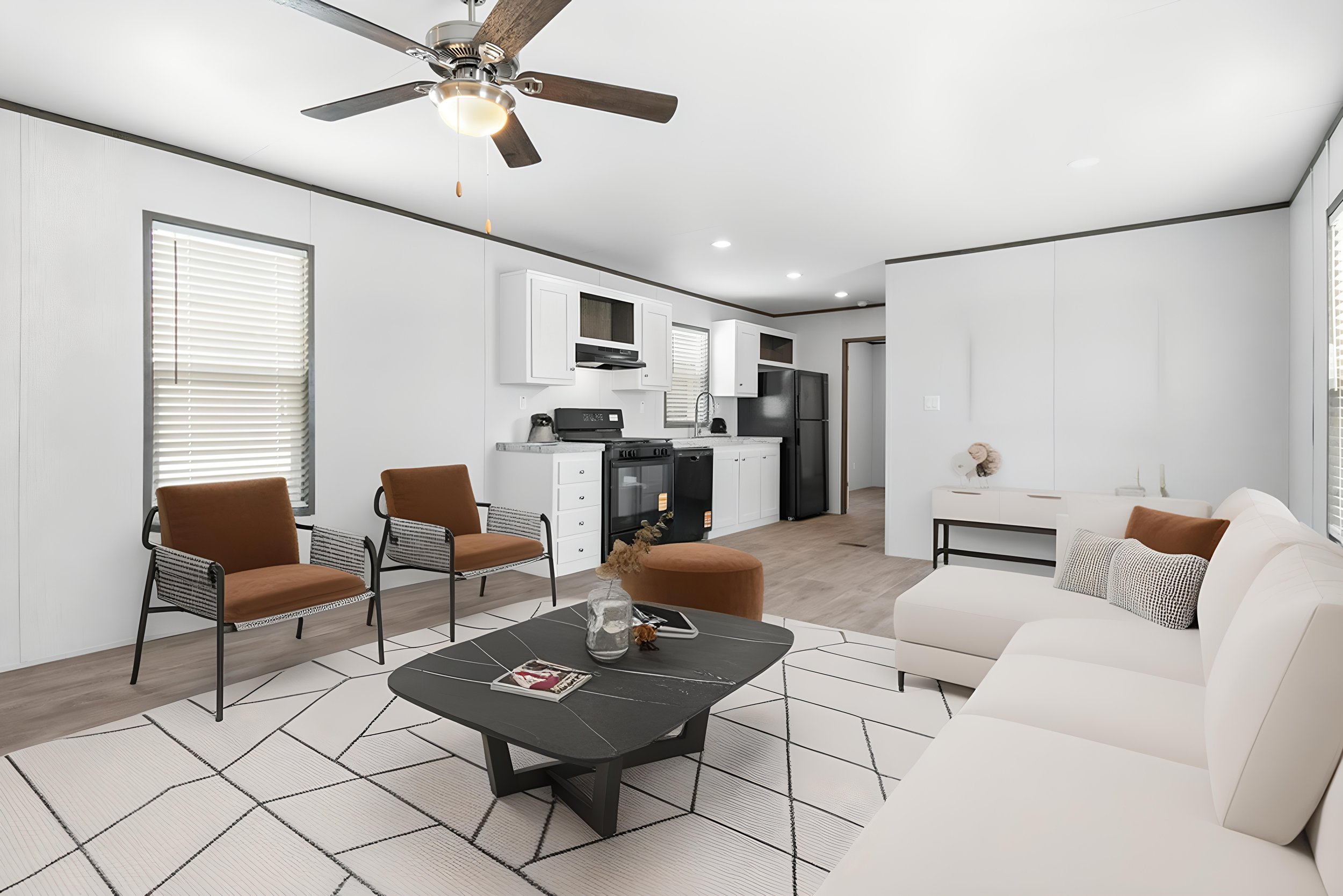 Modern open-concept living room with a white sofa, orange chairs, black coffee table on a white area rug, and a kitchen with white cabinets and stainless steel appliances in the background. Ceiling fan with lights is visible on the ceiling.