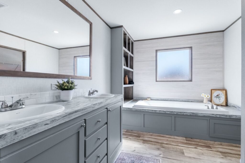A modern bathroom with a large mirror over a marble countertop, a double sink, a potted plant, a built-in bathtub next to a window, a clock, and decorative items.
