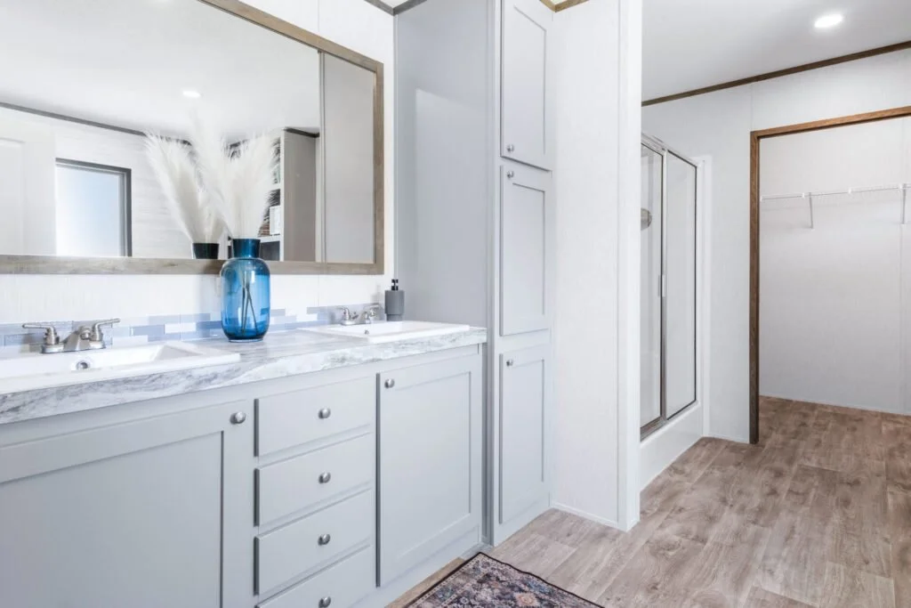 Bathroom with dual sinks, large mirror, white cabinetry, blue vase with dried plants, shower stall with glass door, wooden trim, and beige wood flooring.