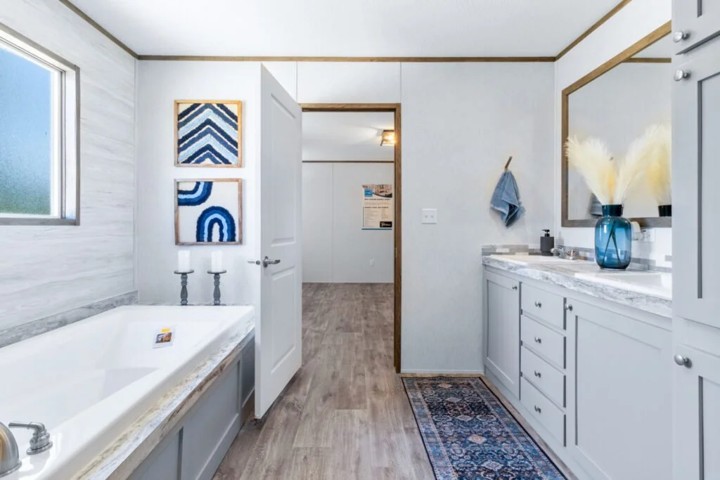 Bathroom with a bathtub on the left, with a window above it, and a vanity with a mirror on the right. Decor includes abstract blue and white artwork, a blue vase with pampas grass, and a blue towel hanging on the wall.