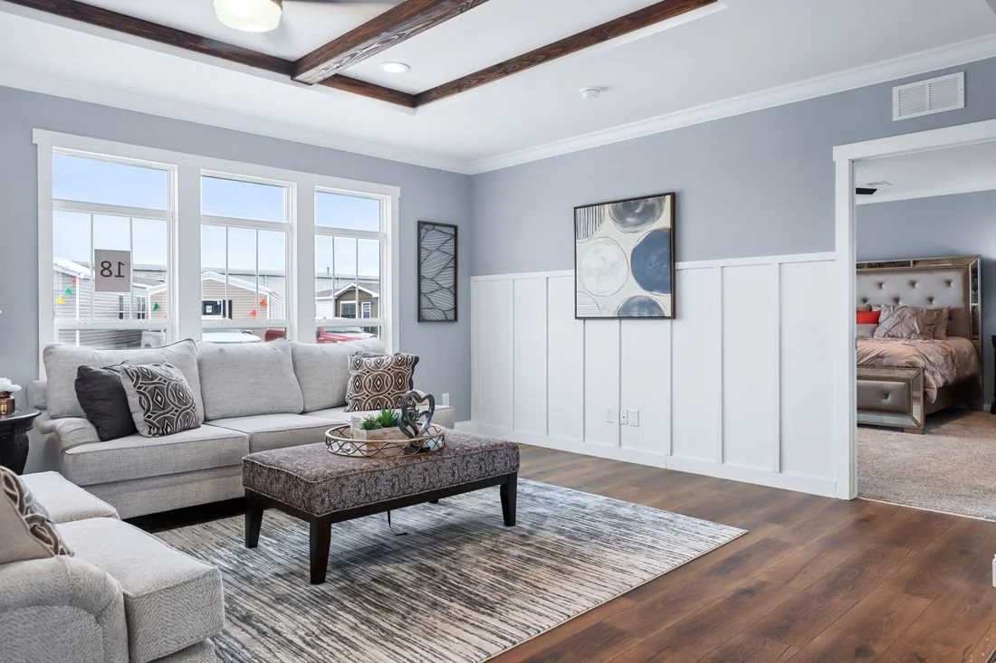 Living room with light gray walls, white wainscoting, wood beam ceiling, hardwood floors, beige sofas, patterned pillows, abstract wall art, windows, and a doorway leading to a bedroom.