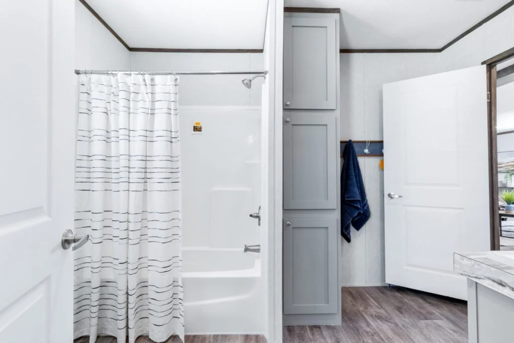 Bathroom with a shower covered by a white curtain with horizontal stripes, gray cabinets, white walls, a door slightly open, and wood flooring.