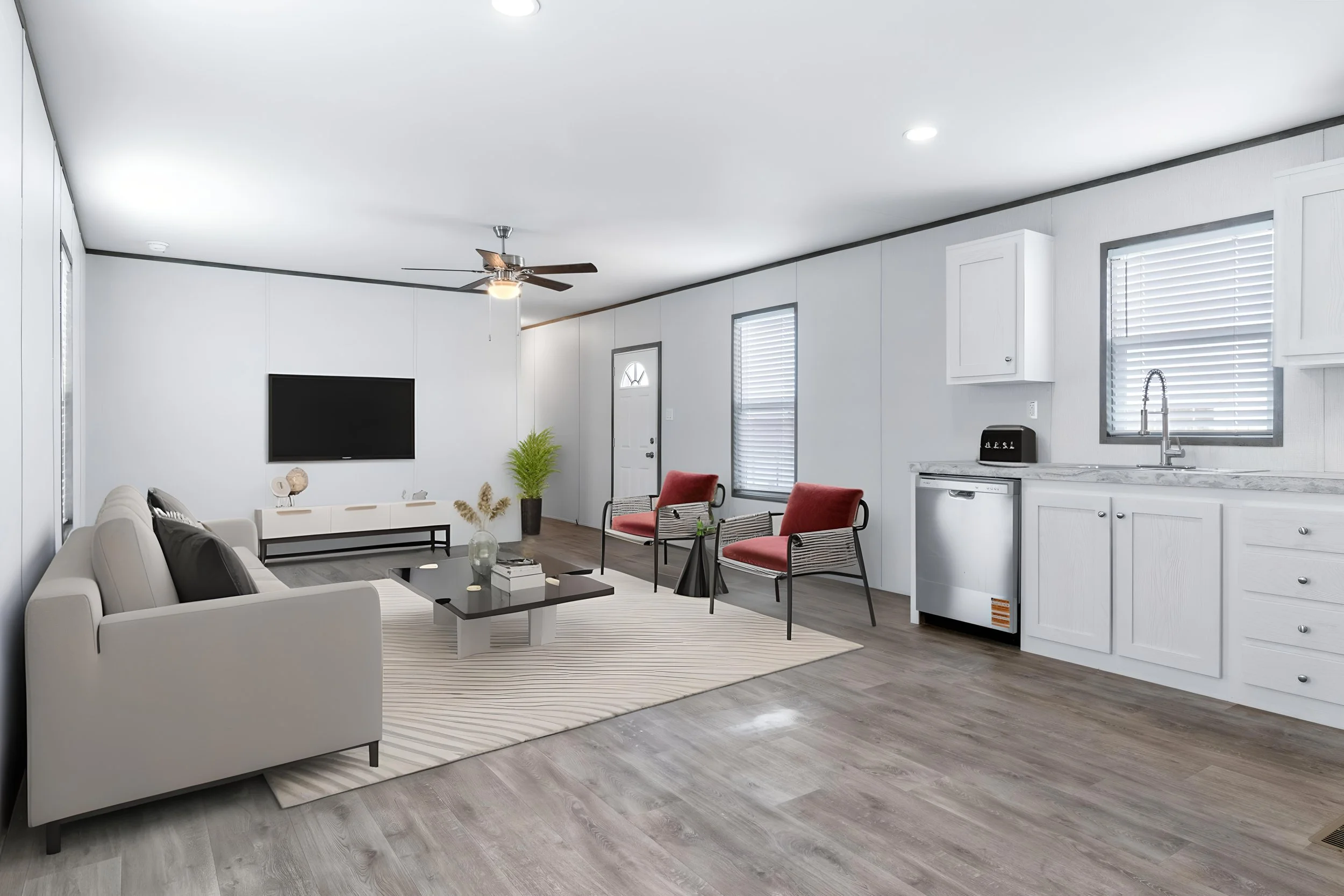 Living room with white walls, a beige sofa with dark pillows, a black coffee table, a striped rug, two red chairs, a wall-mounted TV, and a kitchen area with white cabinets and gray countertops. There are large windows with blinds and a ceiling fan.