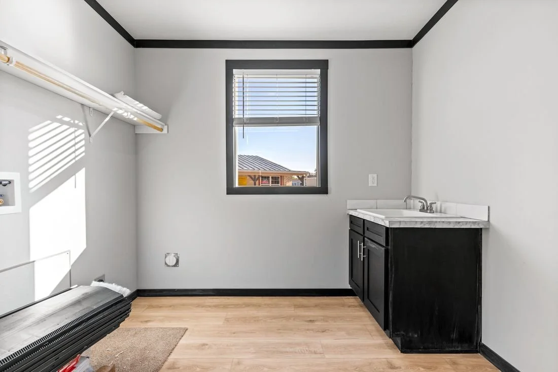 Empty laundry room with white walls, a window, a small black cabinet with a white countertop and sink, and a laundry area with a vent and electrical outlet.