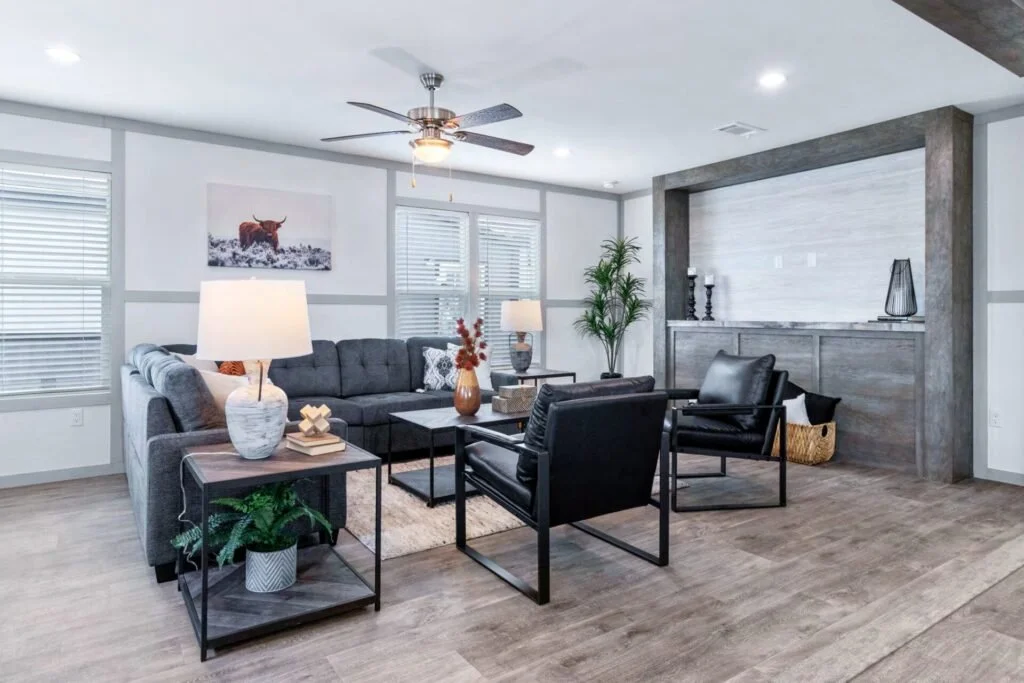Modern living room with gray sectional sofa, black armchairs, wooden flooring, large windows, ceiling fan, decorative items, and a painted cow on the wall.