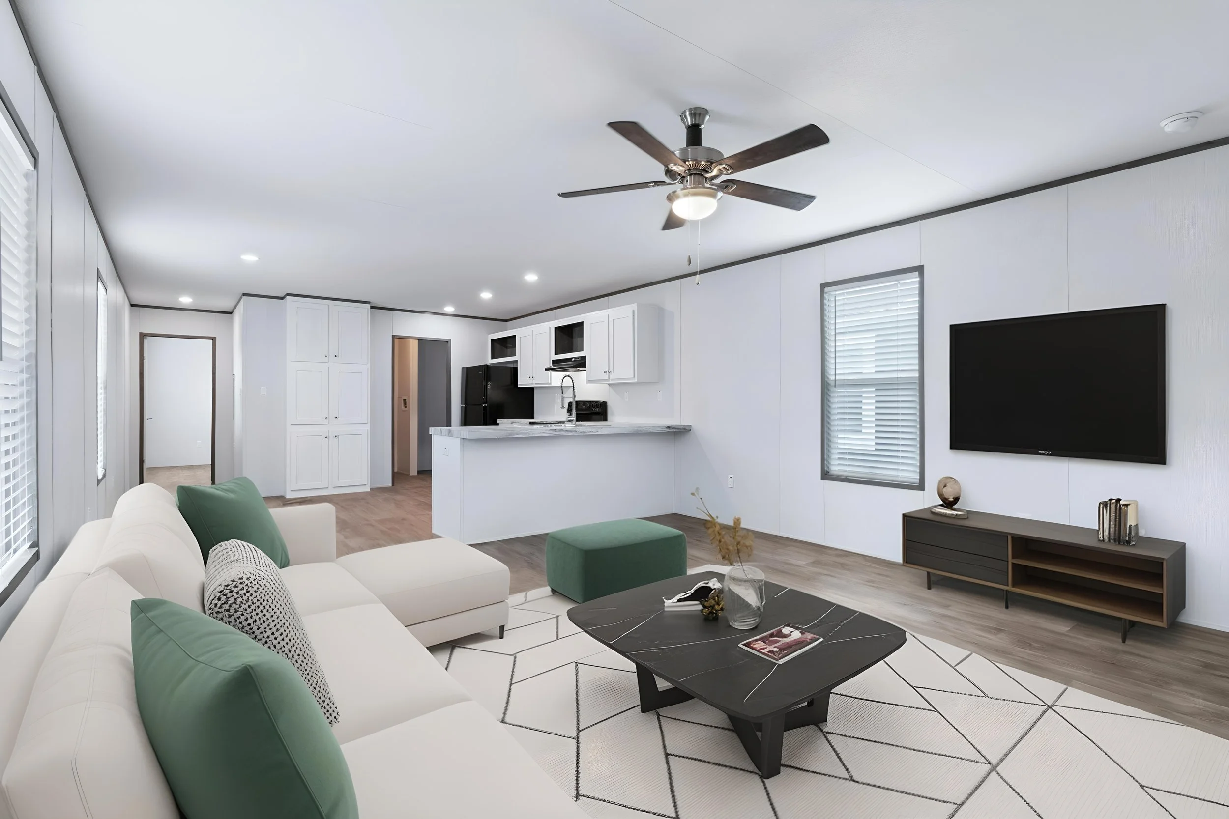 Modern living room with white sofa and green cushions, black coffee table with decor, and a patterned rug. Open kitchen with white cabinets, black appliances, and a marble island. Flat-screen TV on a dark console, and windows with blinds.