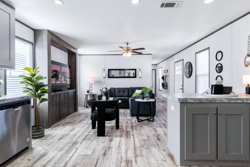 Living room with black sofa, armchair, TV, and modern decor, open to kitchen with gray cabinets and wooden flooring