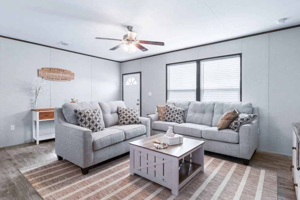 Living room with gray walls, two gray sofas with patterned throw pillows, a wooden coffee table, a window with blinds, a ceiling fan, a small side table with decor, and a doormat near the door.
