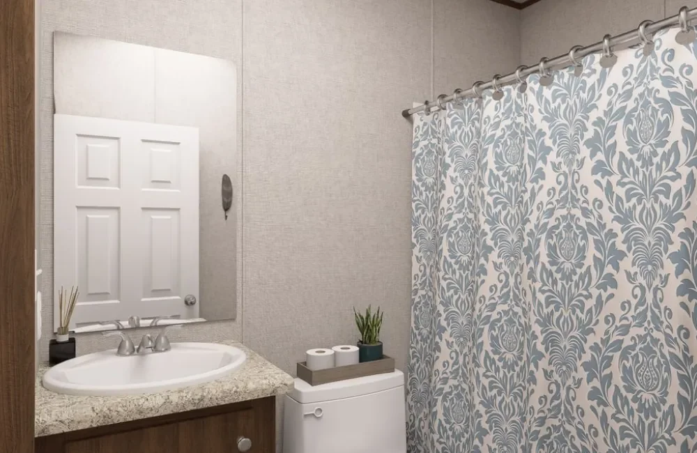 A bathroom with a granite countertop, sink, mirror, shower curtain with blue and white floral pattern, and a small plant on top of a toilet.