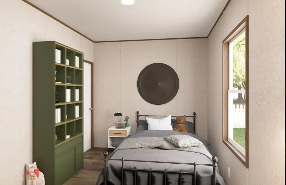 A bedroom with beige walls, a metal bed with gray bedding, a teddy bear on the bed, a small white nightstand with a black lamp, a green bookshelf, and a window showing greenery outside.
