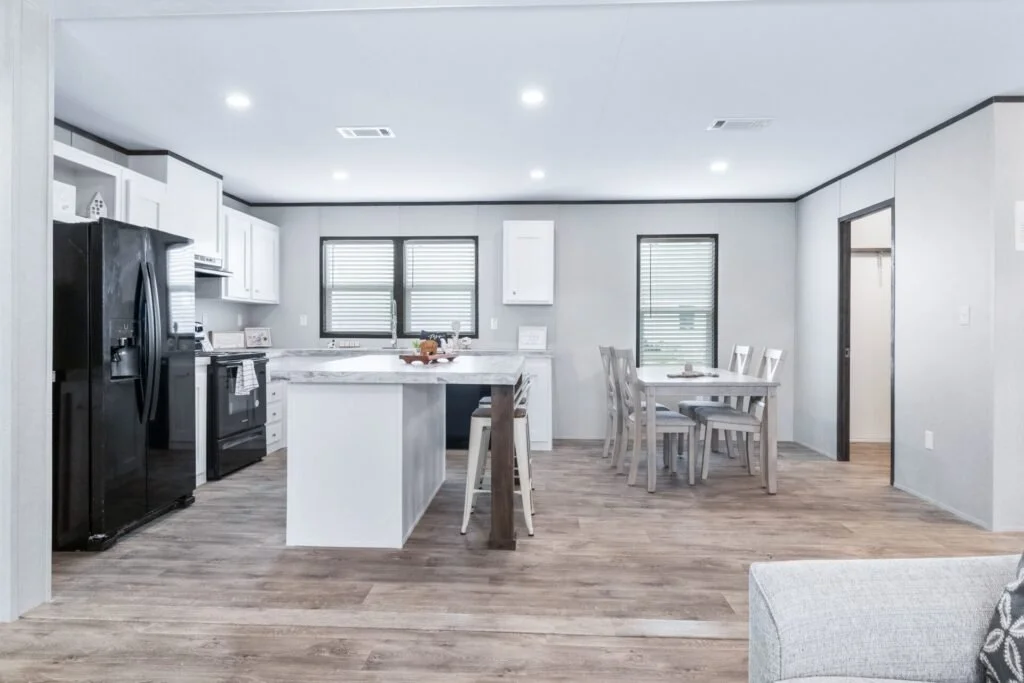Open-concept kitchen and dining area with white cabinets, black appliances, a kitchen island, and a dining table with six chairs. Windows with blinds are on the back wall, and the room has wood flooring.