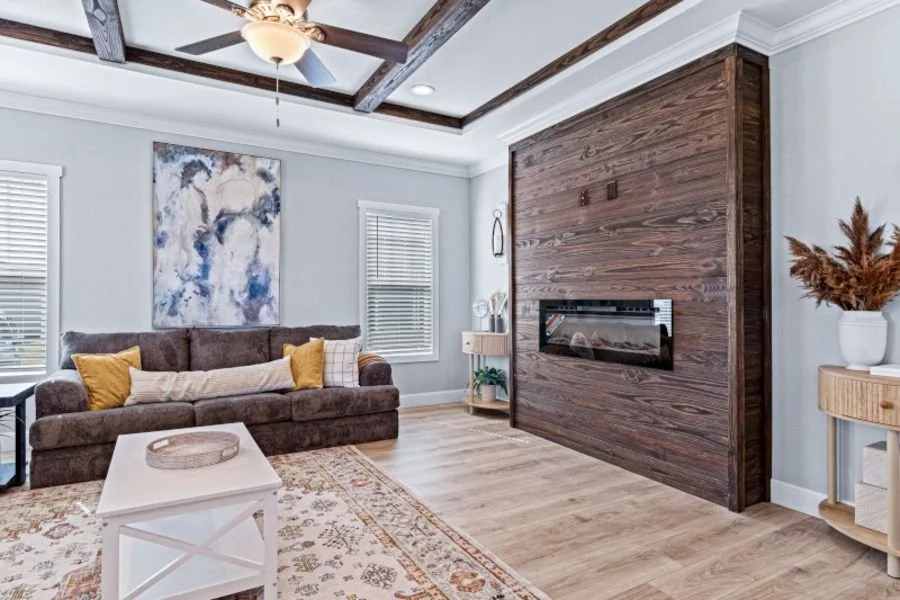 Living room with a brown sofa, yellow pillows, and a white coffee table on a patterned rug. There are two windows with blinds, a large abstract painting on the wall, a modern fireplace built into a wooden wall panel, and a ceiling fan with a light fi