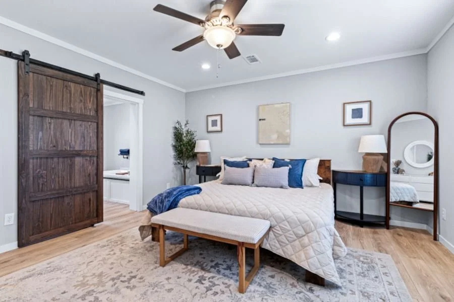A bedroom with a bed featuring a quilted beige cover and multiple pillows, a bench at the foot of the bed, a large mirror on the side, and a ceiling fan with a light. The room has light gray walls, wooden flooring, and wall art.