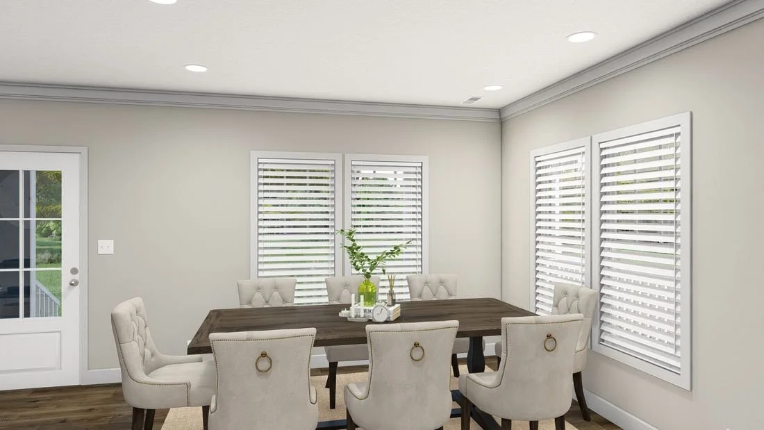 A dining room with beige walls, four windows with white blinds, a dark wooden table, six beige upholstered chairs with nailhead trim, decorative lamps, and indoor plants.