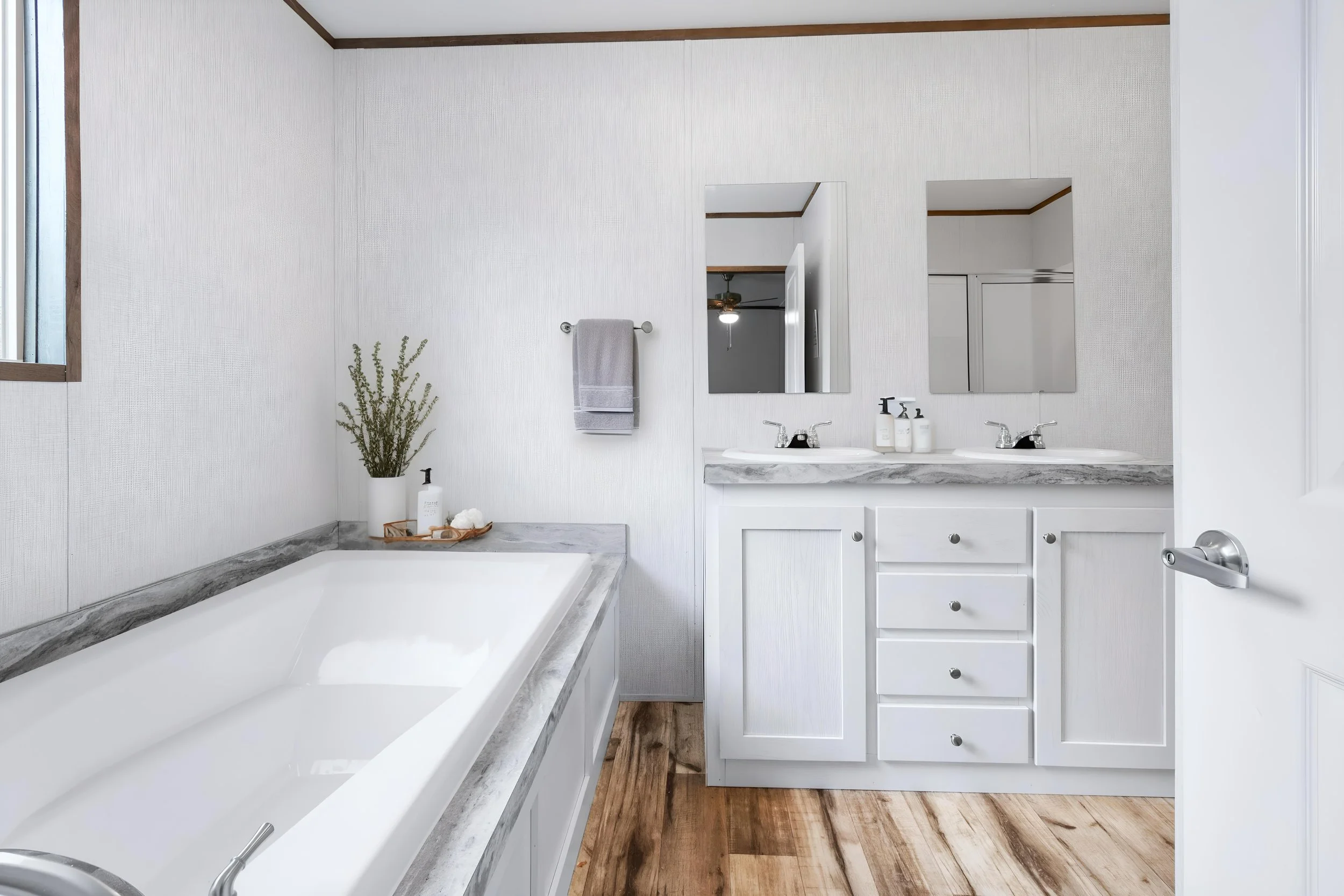 A bathroom with a bathtub, a double vanity with two mirrors, and a window. The bathroom has white cabinets, a gray marble countertop, and wood flooring.