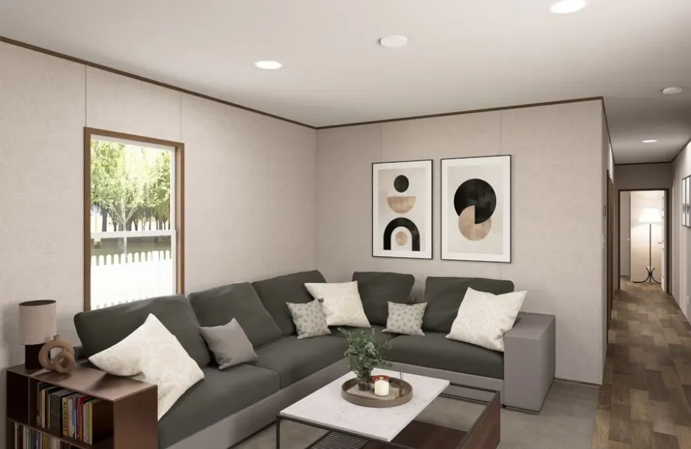 Living room with a gray sectional sofa, framed abstract artwork on the beige wall, small side table with a lamp and books, a window with green trees outside, and a hallway with carpet and a standing lamp in the background.