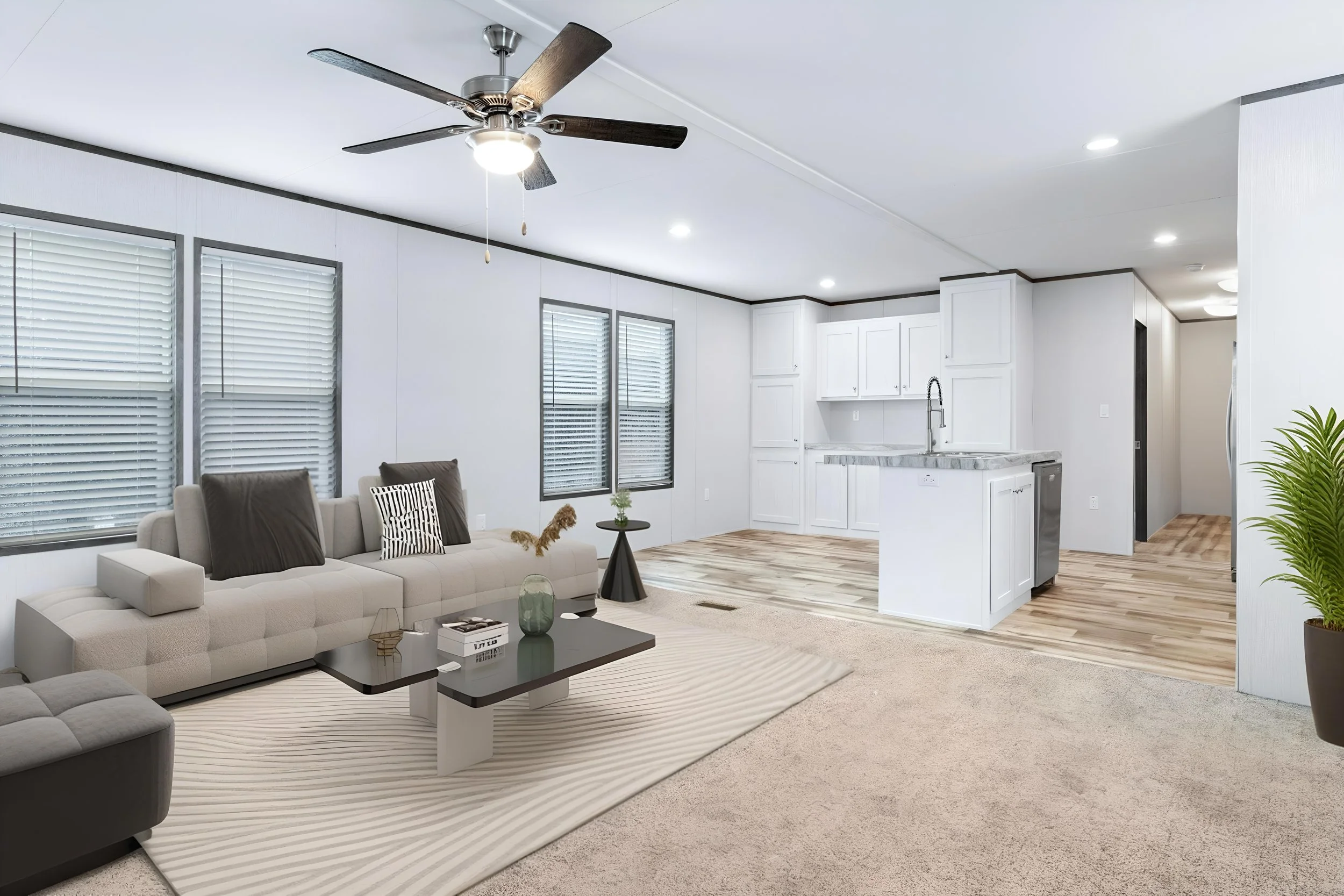 A modern, open-concept living room and kitchen area with white walls, multiple windows with blinds, a ceiling fan with light, a beige sofa with dark pillows, a black coffee table with decorative items, a striped rug, and a kitchen with white cabinetr