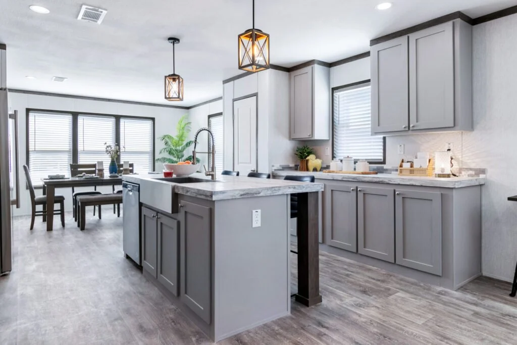 Modern kitchen with gray cabinets, marble countertops, island with sink, pendant lighting, and dining area with large windows and wooden table and chairs.