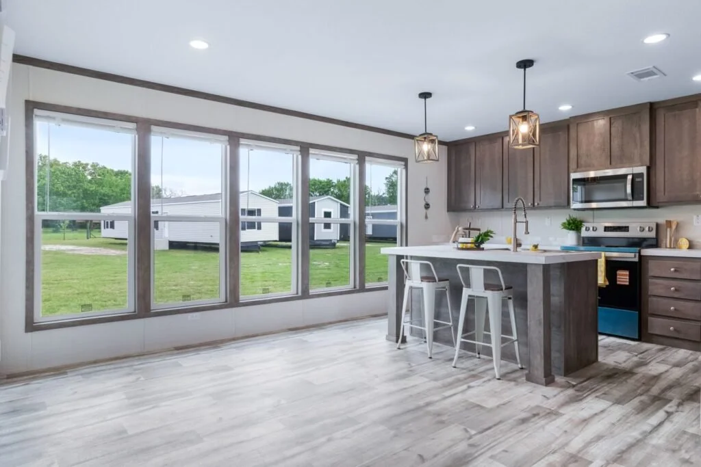 Modern kitchen with large windows, a kitchen island with two white bar stools, pendant lights, wooden cabinets, and stainless steel appliances.