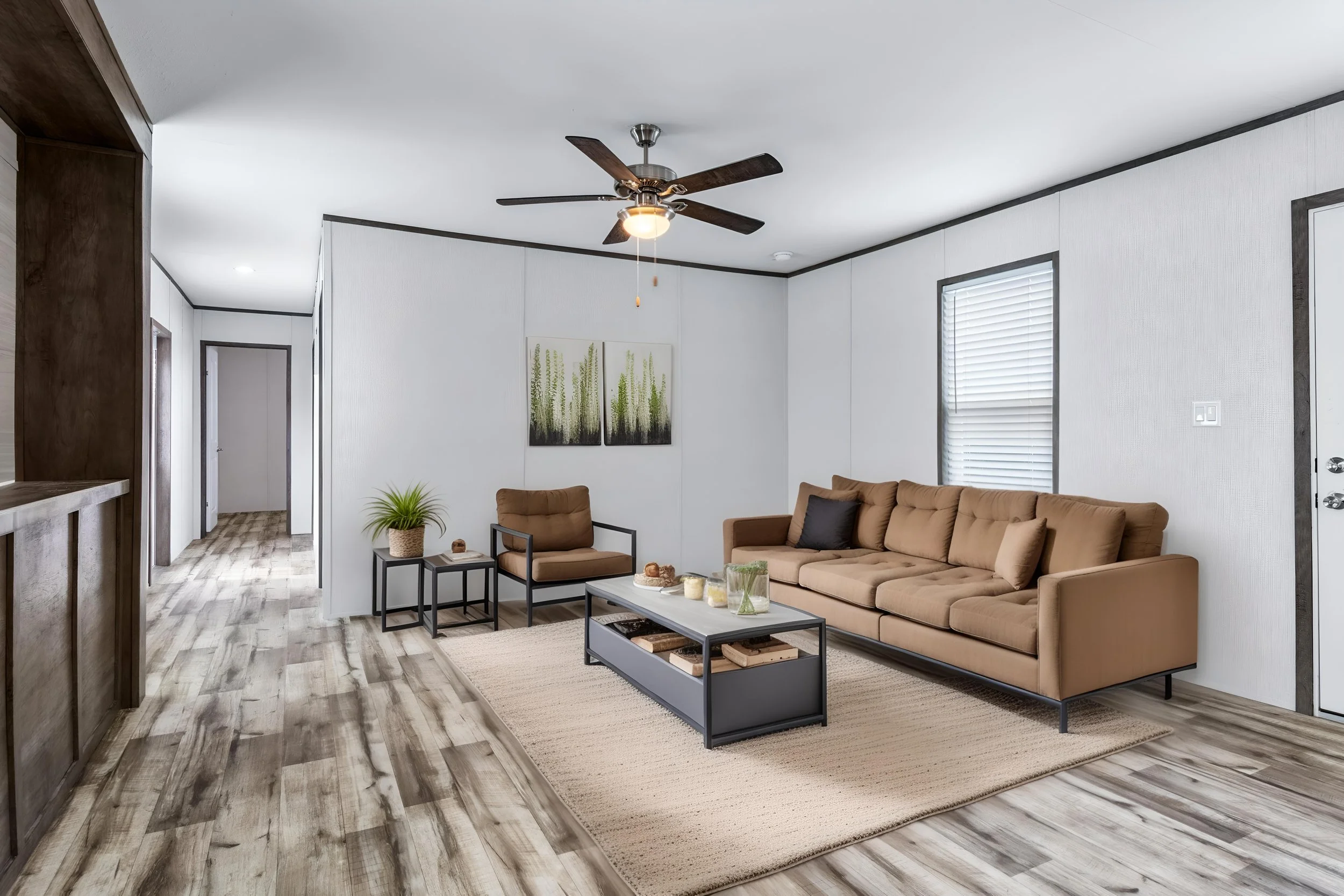 Living room with beige couch, two brown armchairs, coffee table, framed art of green plants, window with blinds, ceiling fan, wood flooring, white textured walls, and a beige rug.