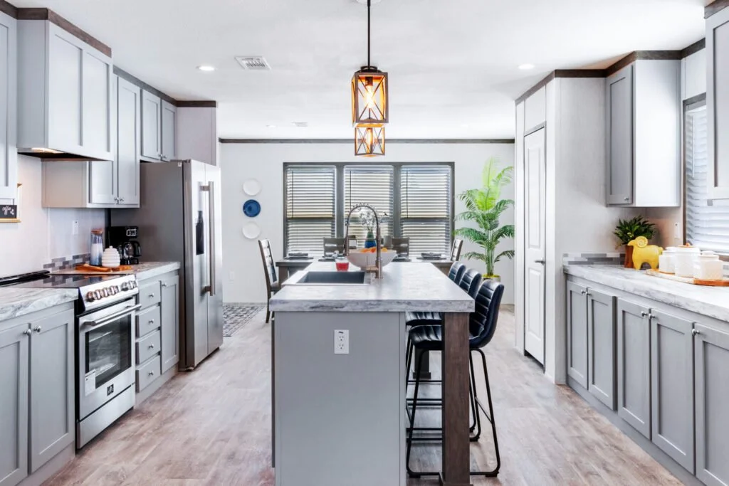 Modern kitchen with gray cabinets, white marble countertops, stainless steel appliances, a kitchen island with barstools, and a dining area with a large window and green plant.