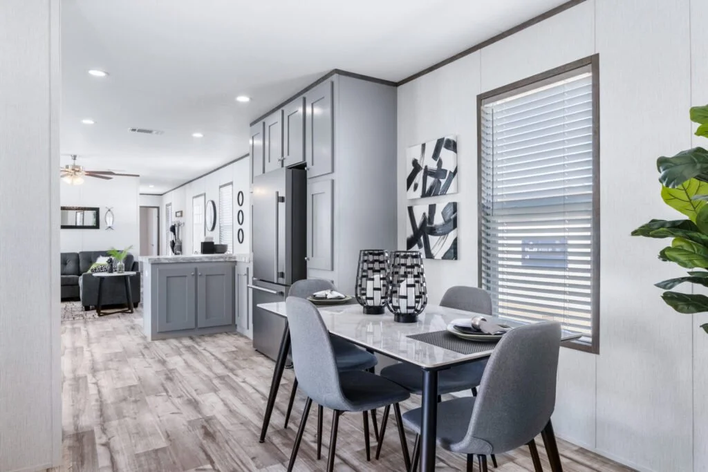 Dining area with a rectangular table, four gray chairs, black and white vases, and wall art, adjacent to a kitchen with gray cabinets and stainless steel refrigerator, in a modern open-plan home.
