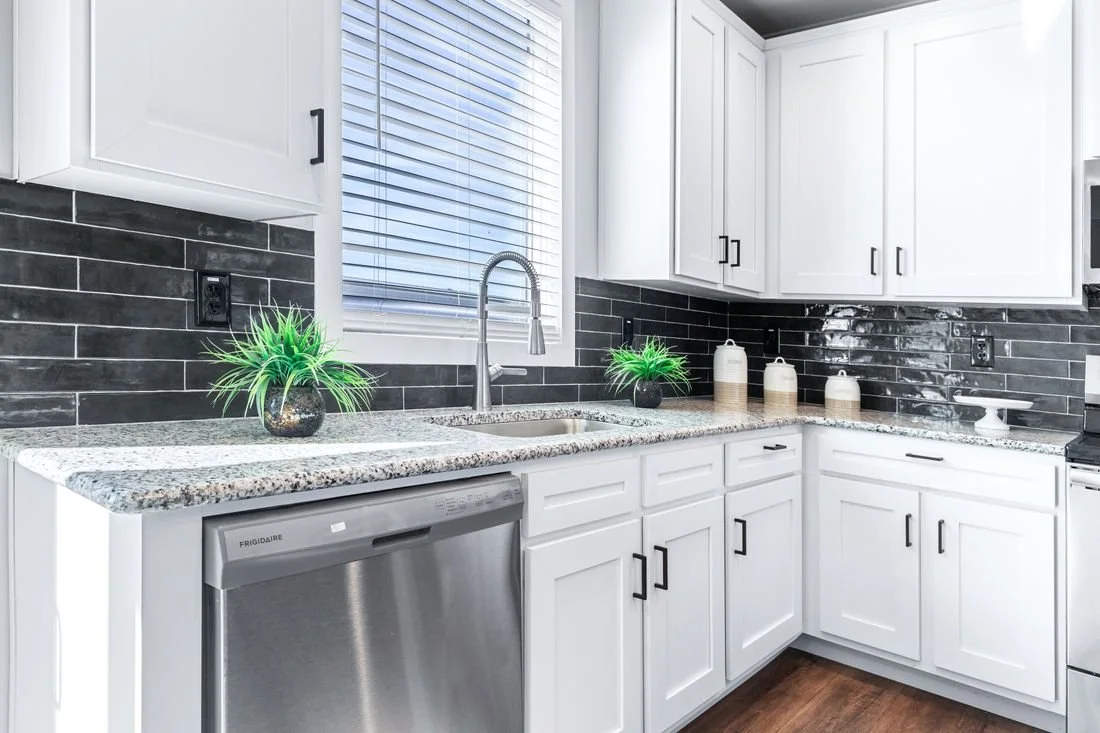Kitchen with white cabinets, black tile backsplash, granite countertops, a stainless steel dishwasher, a window with blinds, potted plants, and decorative jars.