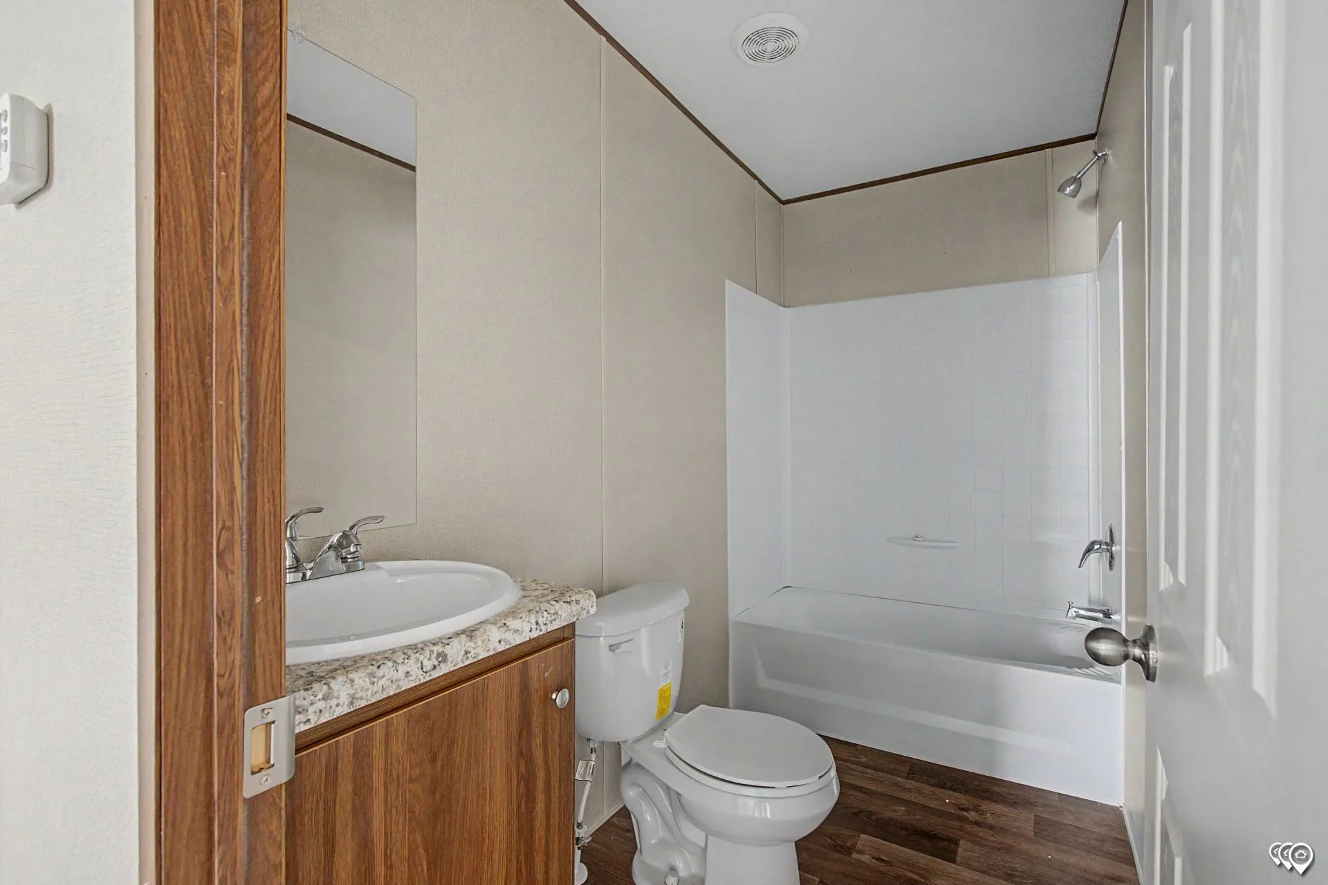 Small bathroom with a white bathtub, a toilet, a wooden vanity with a white sink, a granite countertop, and a mirror. The walls are covered with beige wallpaper, and the floor is dark brown wood. There is a small light fixture above the bathtub.