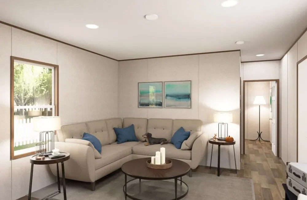 Living room with white sectional sofa, blue pillows, wooden side tables with lamps, wall art, window with green trees outside, and a small dog resting on the sofa.