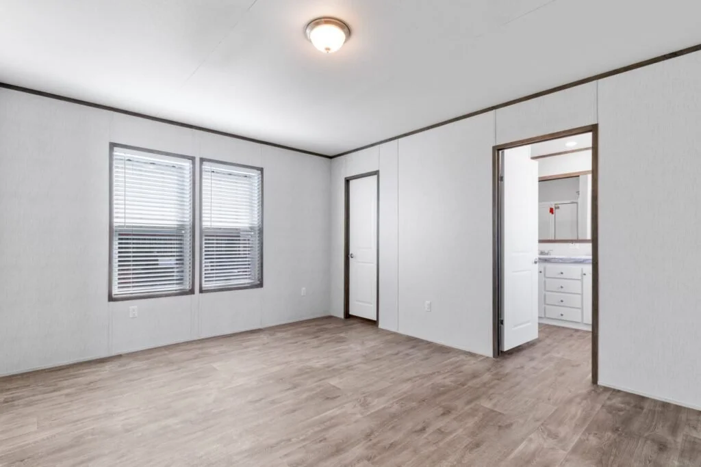 Empty room with white walls, wood flooring, two windows with blinds, a door, and a view of a white bathroom with cabinets.