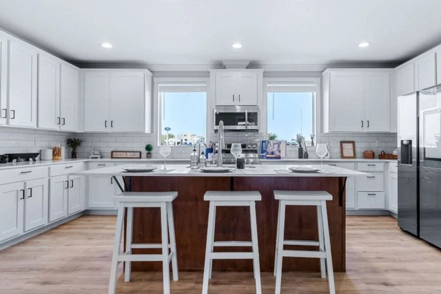 Modern white kitchen with island, three white stools, stainless steel refrigerator, built-in oven, and two windows
