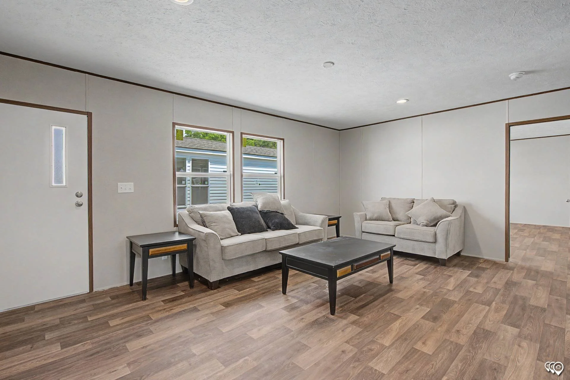 The image shows a living room with a white sofa, a matching loveseat, and two black side tables with yellow accents. There are several throw pillows on the sofa and loveseat. The room has two windows and a wooden floor.