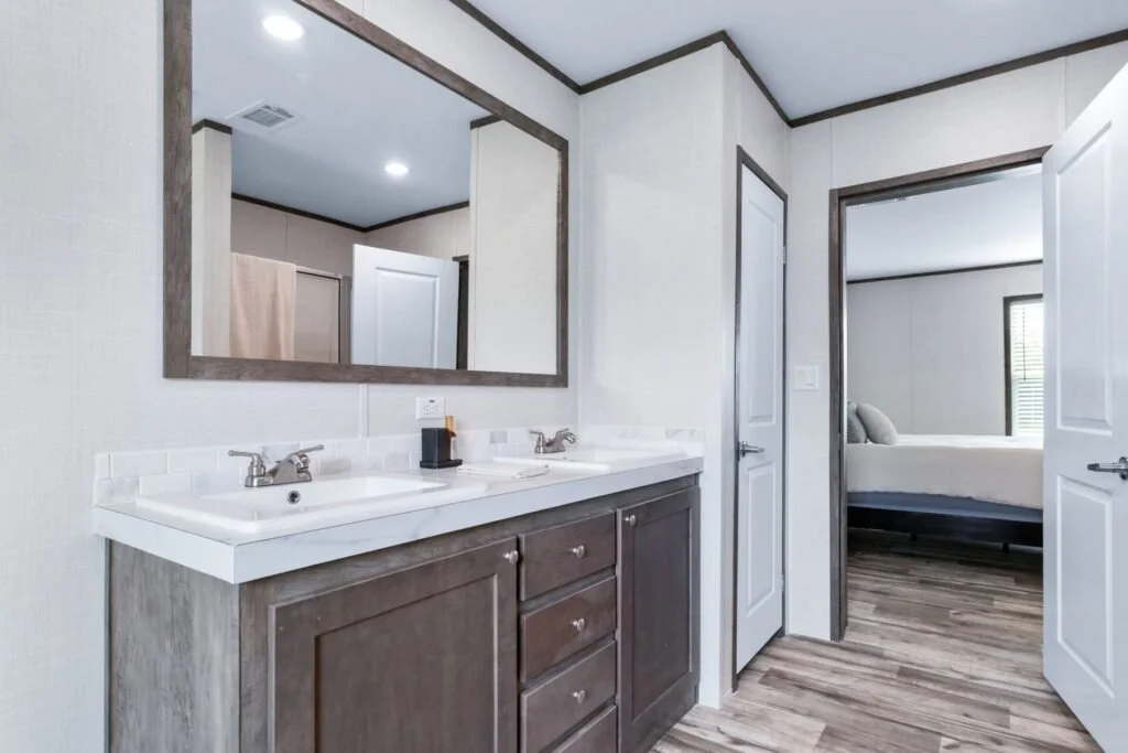 Bathroom with dual sinks, large mirror, and door leading to bedroom with bed and window.