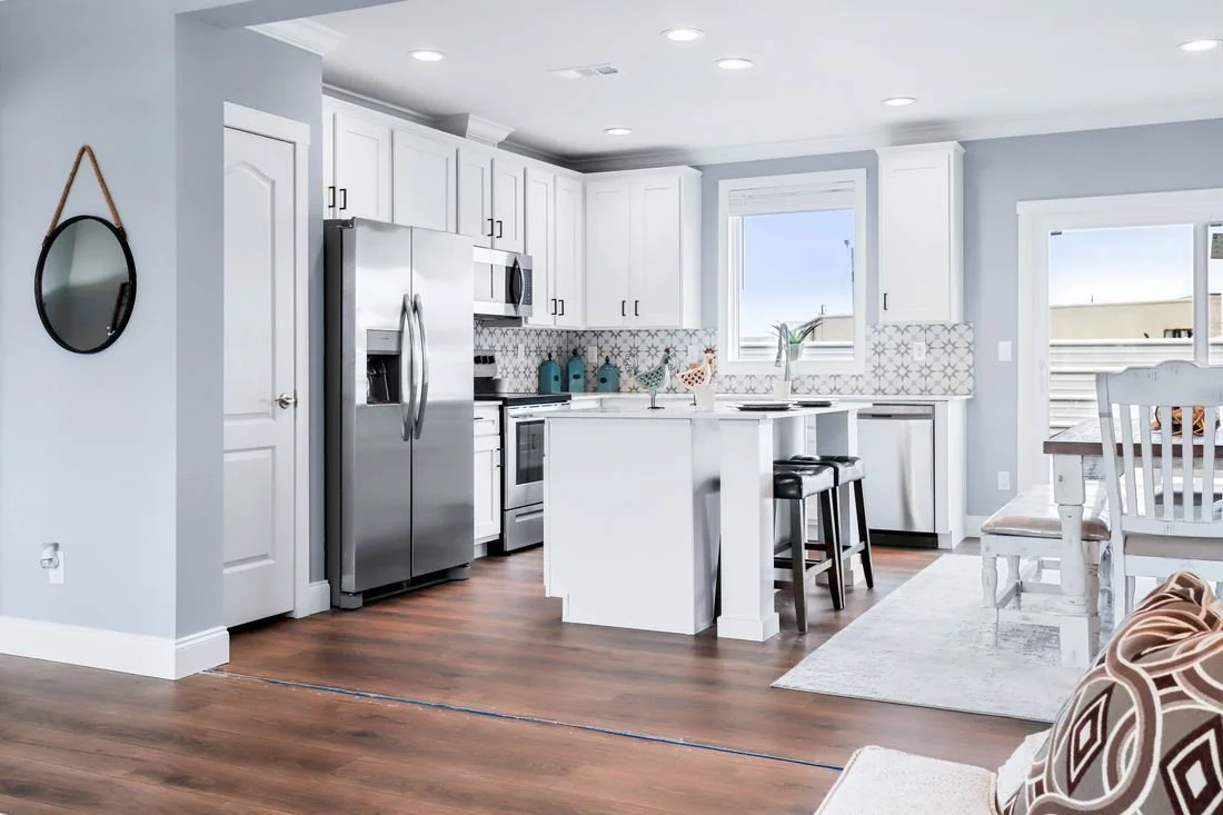 Clean, modern kitchen with white cabinets, stainless steel appliances, wooden flooring, and a small island with a black stool. There is a window above the sink and a sliding glass door leading to outside.