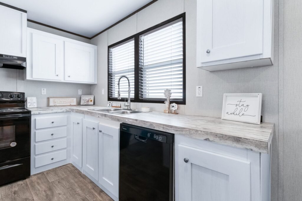 A modern kitchen with white cabinets, a black stove, a black dishwasher, a marble countertop, two large window blinds, and decorative signs including one that says "stay humble."