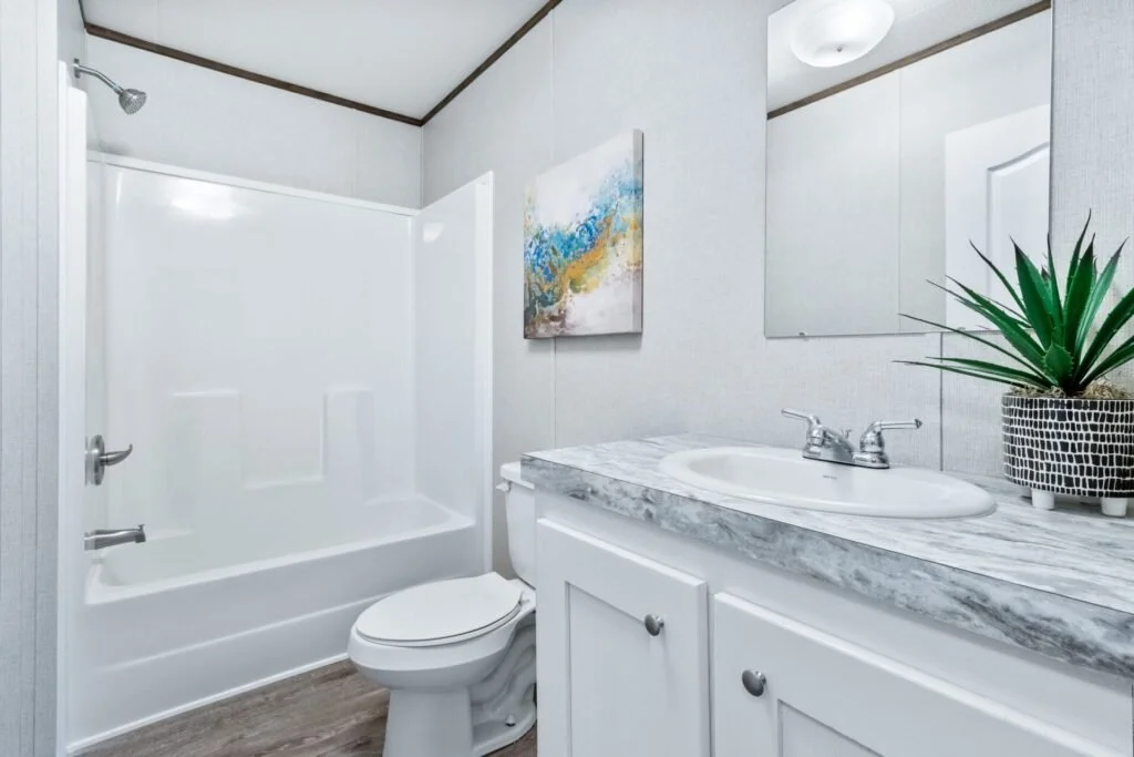 A small bathroom with a bathtub and shower, a toilet, and a vanity with a marble countertop, sink, and mirror. There is a potted plant and a piece of abstract wall art.