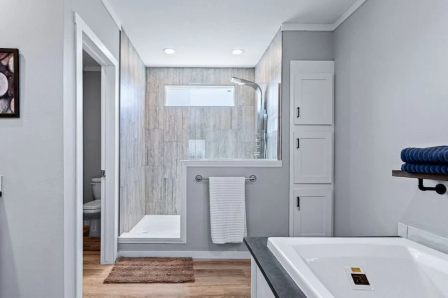 Modern bathroom featuring a walk-in shower with glass enclosure, a toilet in a separate room, a corner whirlpool tub, and white cabinetry, with light gray walls and wood-look flooring.