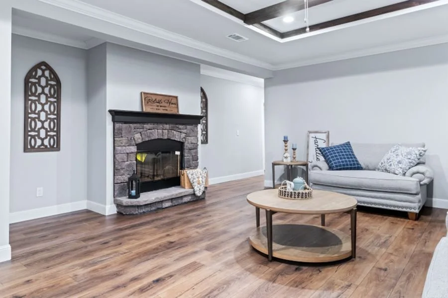 A living room with a gray couch, blue and white pillows, a round wooden coffee table with a decorative tray and tea cups, a fireplace with a stone surround, wooden framed wall decorations on each side of the fireplace, and a wooden side table with ca