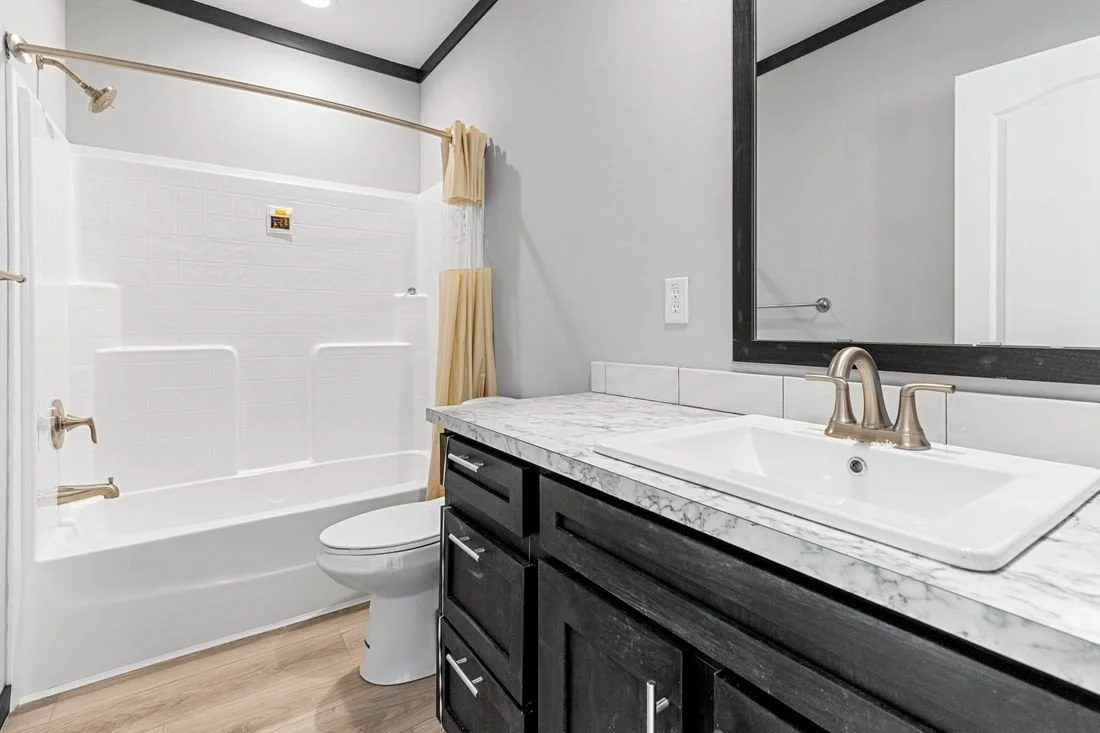 Bathroom with a white bathtub and shower, a toilet, a dark-colored vanity with a marble countertop, a sink with a bronze faucet, and a large mirror.