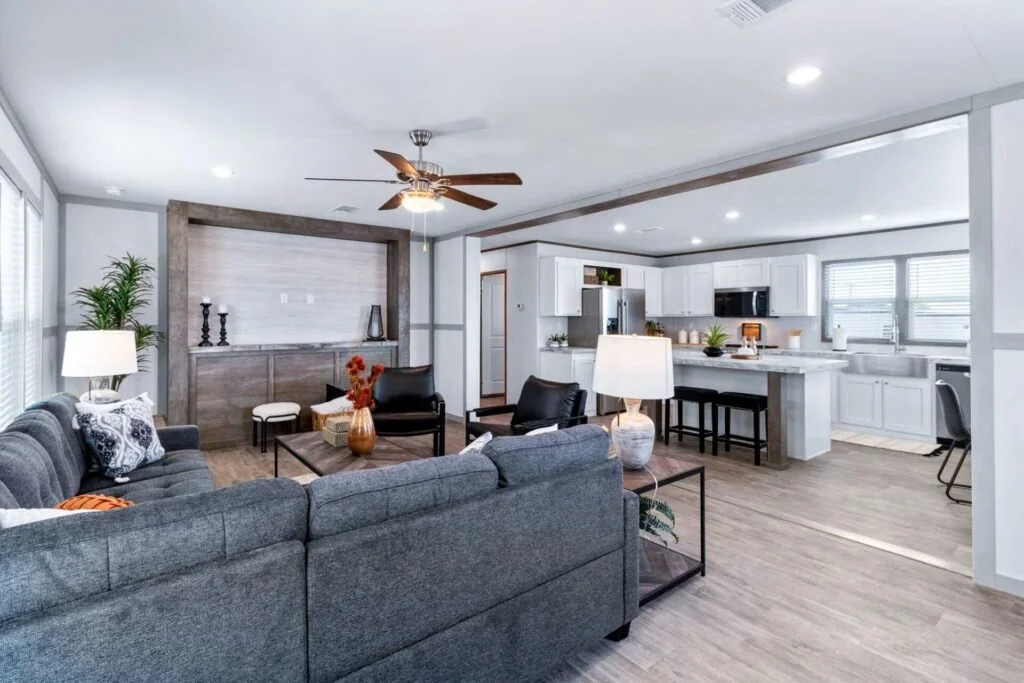 Open-concept living room and kitchen with white cabinets, gray sofa, black chairs, and wood accents, sunlit from multiple windows.