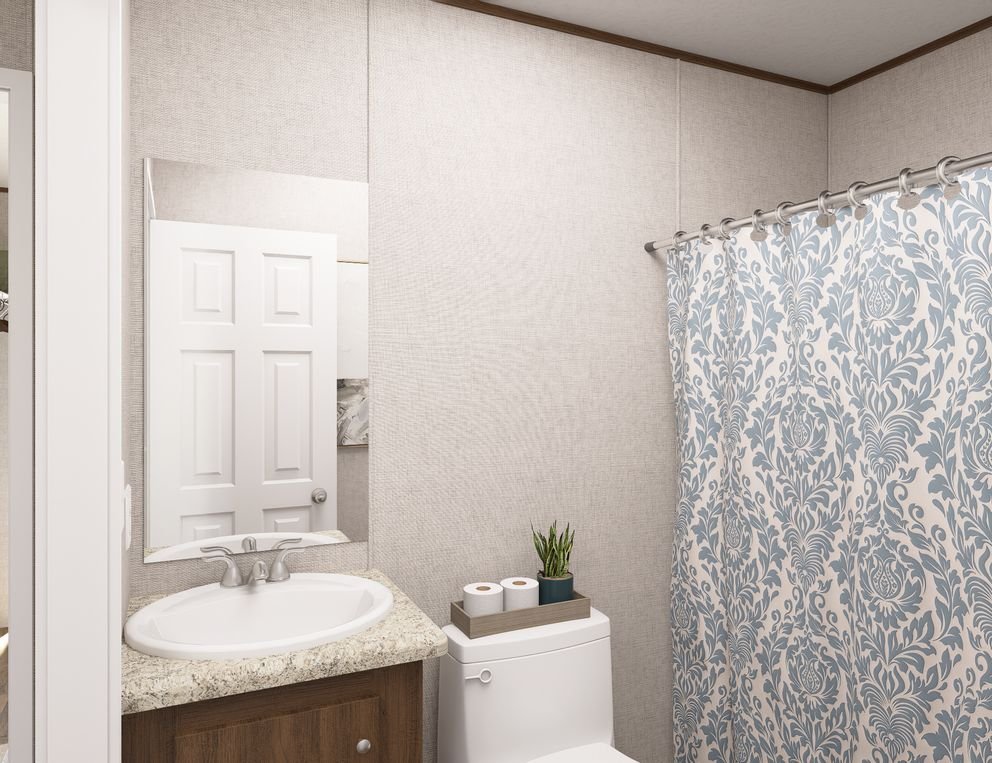 Small bathroom with a granite countertop sink, a mirror, a toilet with a tray holding two rolls of toilet paper and a small plant, and a shower with a patterned blue and white curtain.