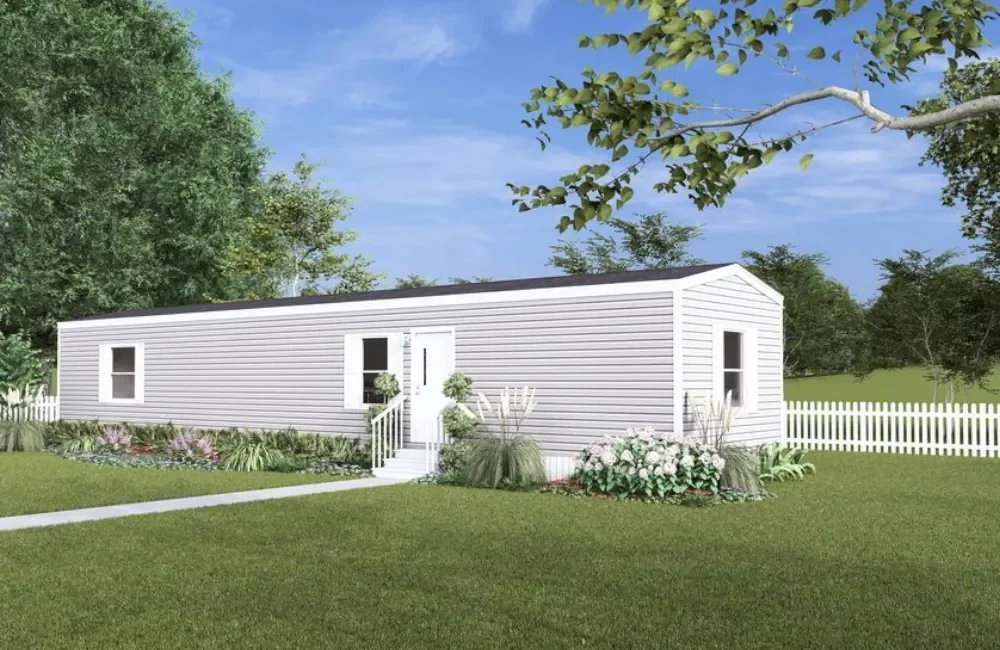 A white manufactured home with black roof shingles, surrounded by green trees, bushes, and a white picket fence, under a blue sky.