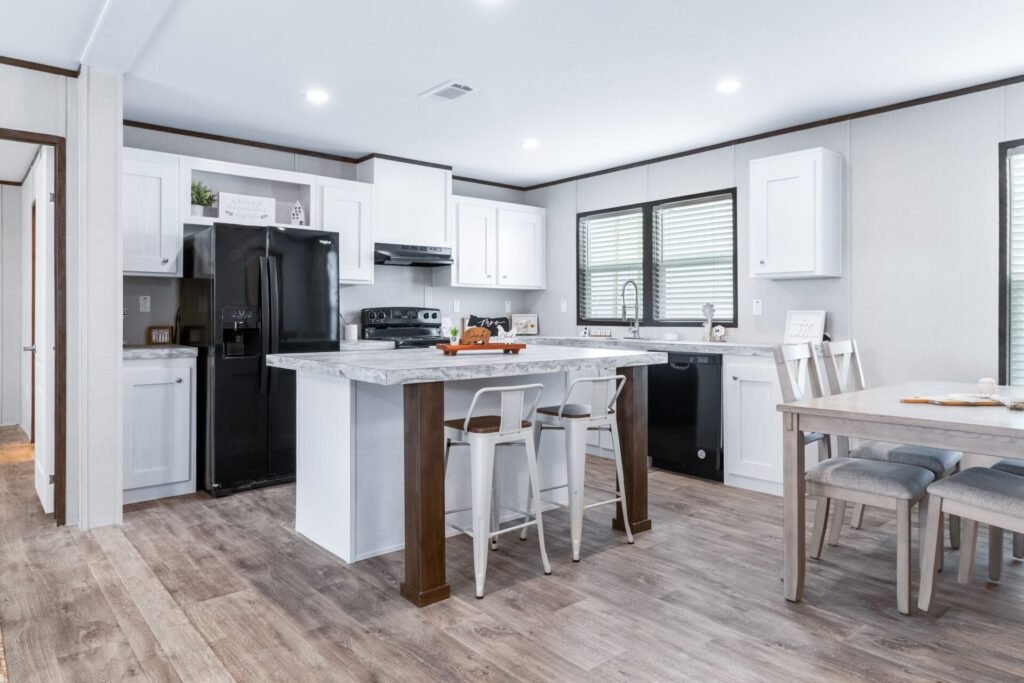 Modern kitchen with white cabinets, black appliances including refrigerator and dishwasher, gray countertops, and wood flooring. There is a kitchen island with chairs, a dining table with chairs, and a window above the sink.
