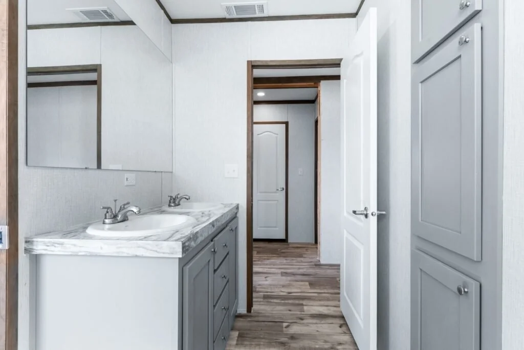 Inside a bathroom with a double sink vanity with a marble countertop, a large mirror, and light-colored cabinetry. There is a door open to the hallway, and a tall cabinet to the right.