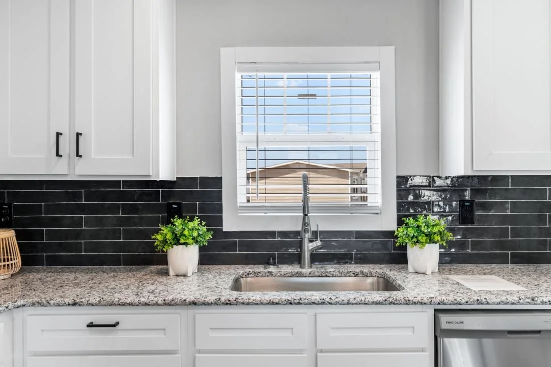 Kitchen window above a sink with two potted plants on a granite countertop, black tile backsplash, white cabinets, and a dishwasher.