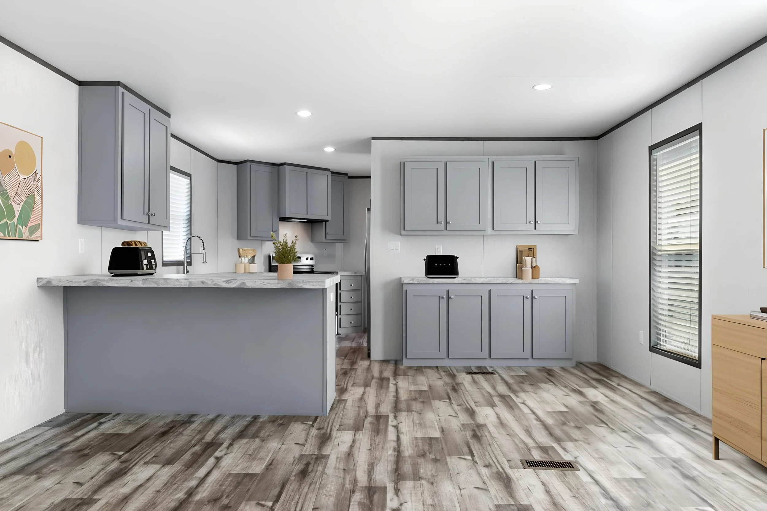 A modern kitchen with gray cabinets, marble countertops, and wood-look flooring. There are windows with blinds, and a small artwork on the wall.