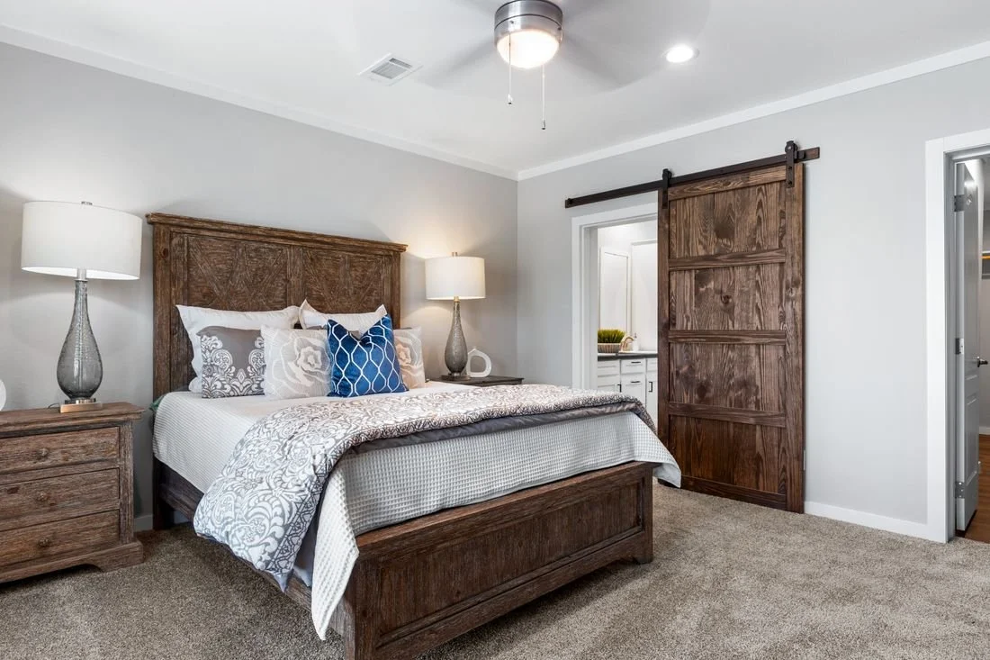 Bedroom with wooden bed frame, nightstands, and sliding barn door, neutral decor, carpeted floor, two lamps, and an open bathroom visible in the background.