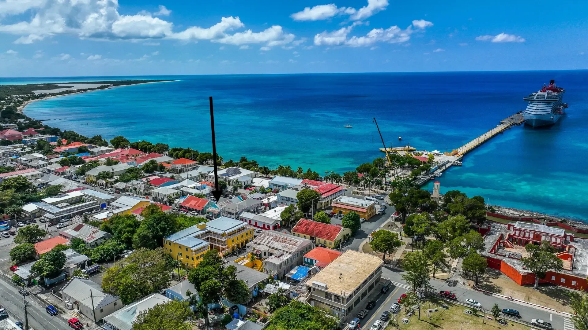 Aerial view of a coastal town with colorful buildings, a pier extending into the blue ocean, a cruise ship docked at the pier, and a sandy beach with clear waters in the background.
