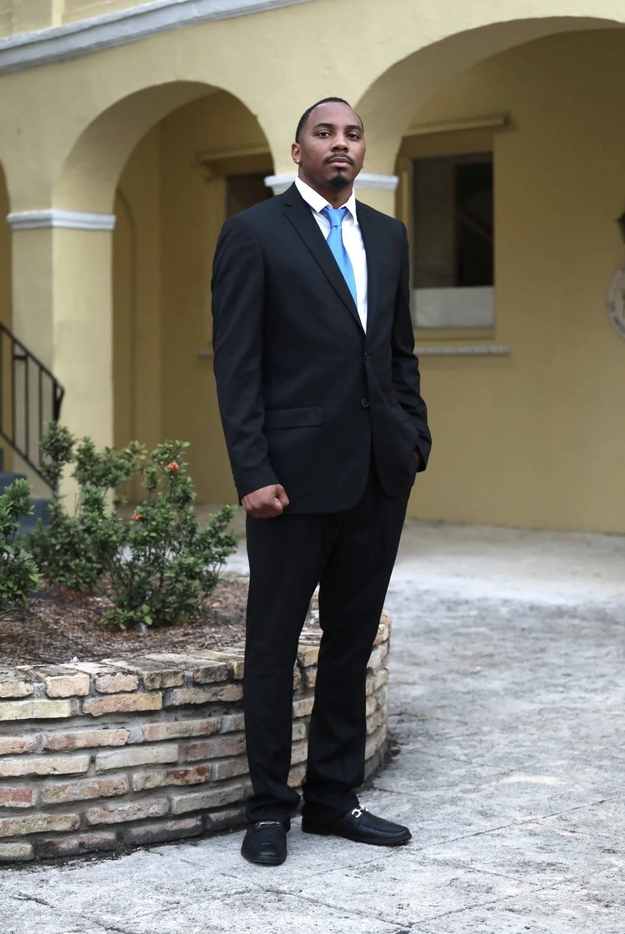 A man in a black suit with a blue tie standing outdoors in front of a yellow building with arches.